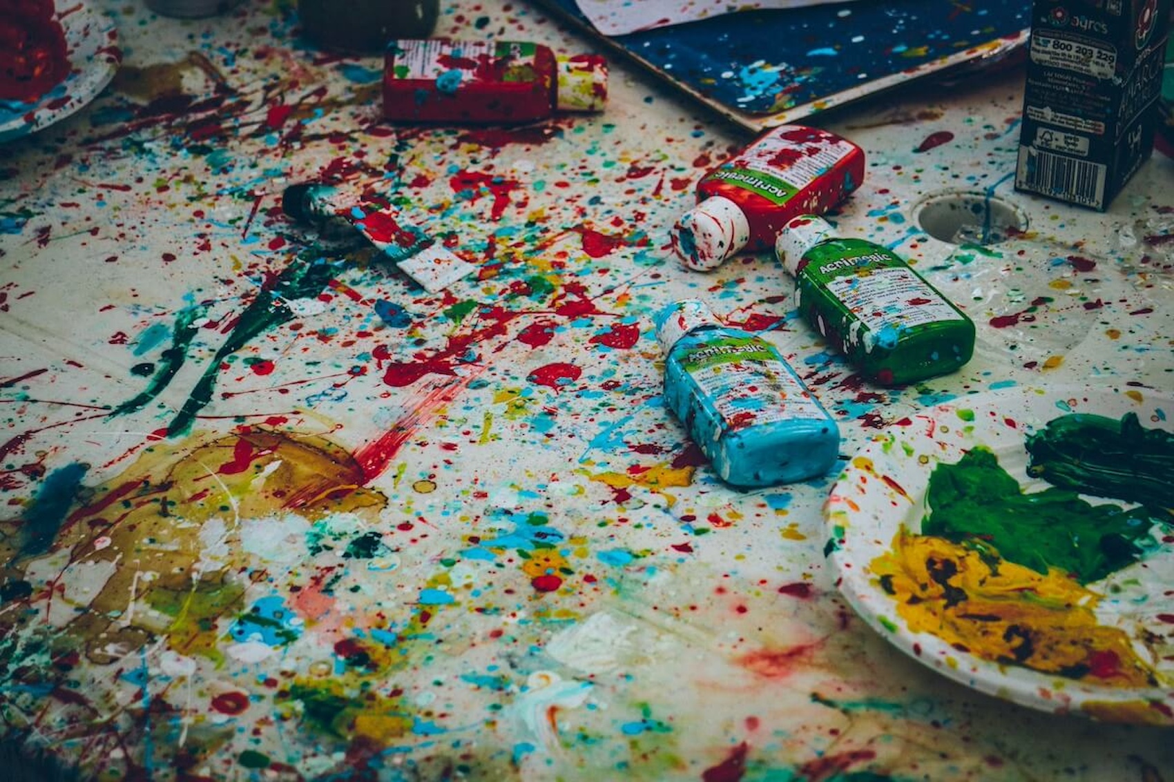 A messy, paint-splattered surface covered in colourful art supplies and vibrant splatters of paint, representing the uninhibited creativity and free-flowing imagination of a child's mind that writers should strive to emulate. This is one of the key tips for writers.