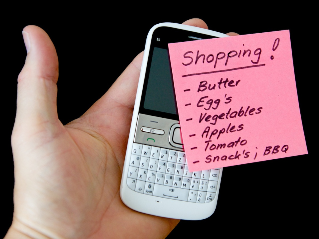 A hand holding a phone with a shopping list, highlighting the incorrect use of an apostrophe in the plural form "egg's".